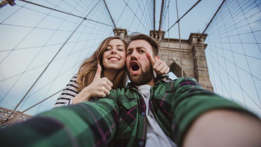 Beautiful happy couple taking selfie self-portrait on Brooklyn Bridge, New York. Hipster tourists having fun and photographing NY landmarks for travel blog.